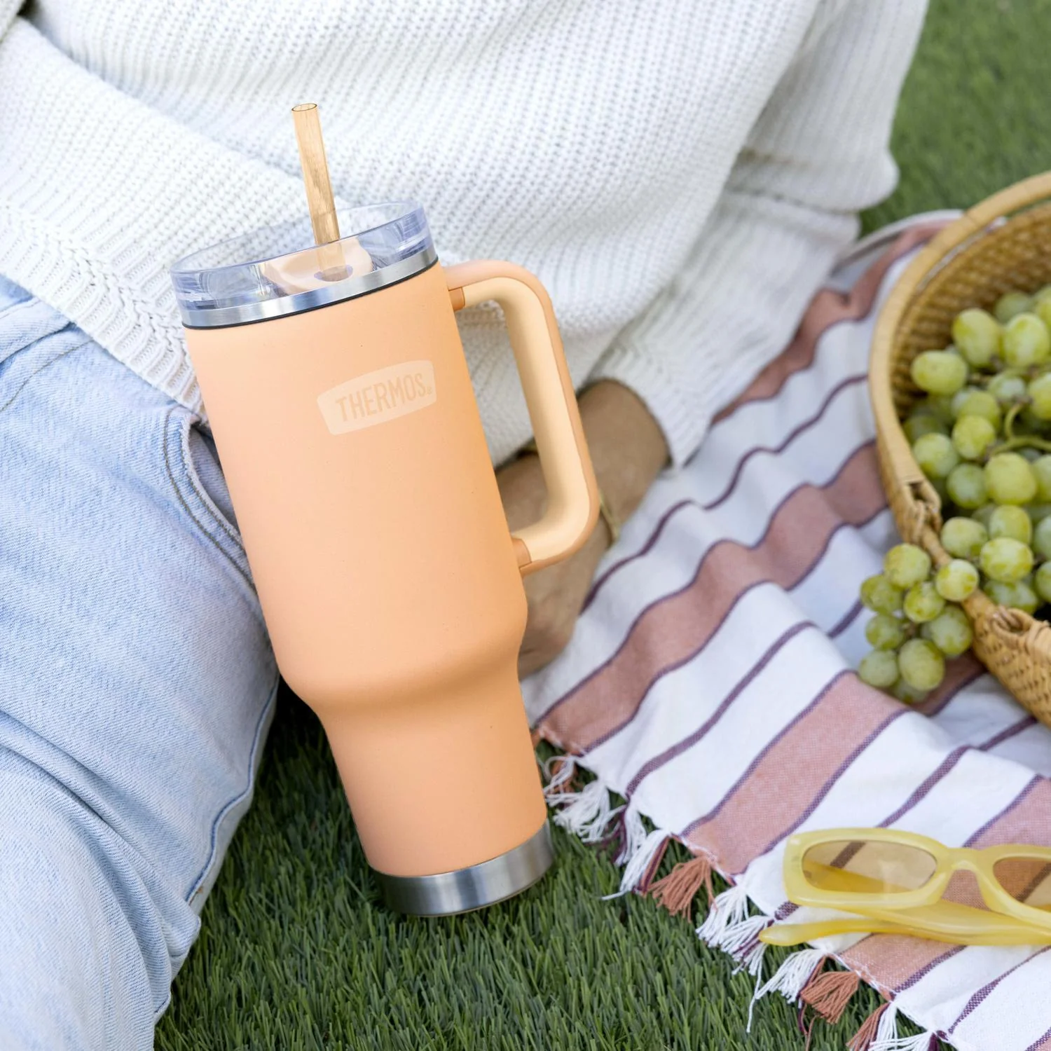 Person holding a peach-colored tumbler with a straw on a picnic blanket with grapes and sunglasses.